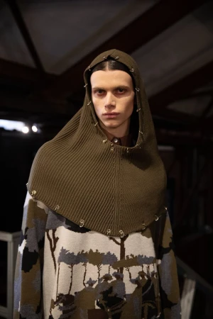 Close-up studio shot of a fashion model wearing an olive green hooded head covering and printed sweater with tree silhouettes.