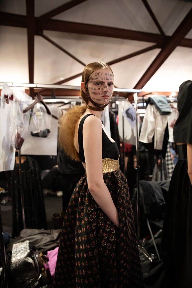 Fashion model with text on face in black and gold dress backstage at a show, indoor lighting, dark background, medium shot.