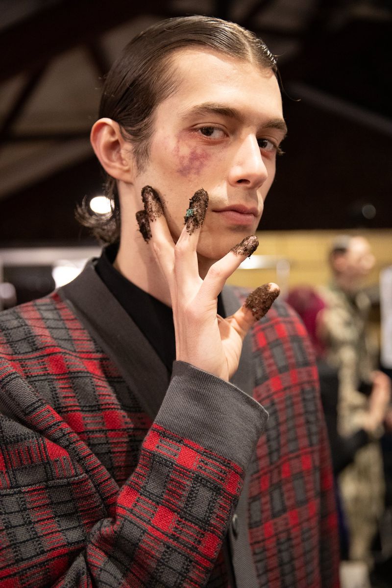 Close-up of a young adult male with slicked-back hair wearing a red and gray plaid jacket and hands with a dark substance on his fingers.