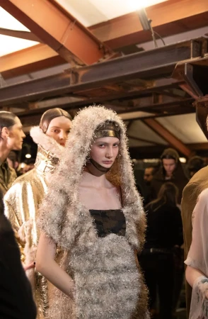 A model in an avant-garde fashion show, wearing a hooded silver dress and metal headpiece, standing among other fashion models.