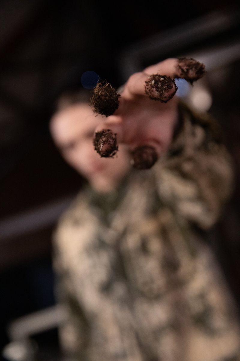 Close-up of an outstretched hand with dirty fingertips; blurred young adult wears camo shirt against a dark background.