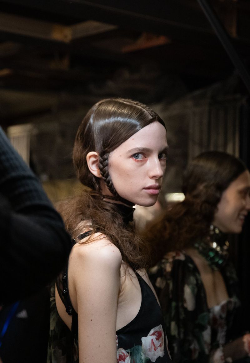 Portrait of a fair-skinned woman with brown hair styled in braids, wearing a floral top, and a choker necklace in an indoor setting.