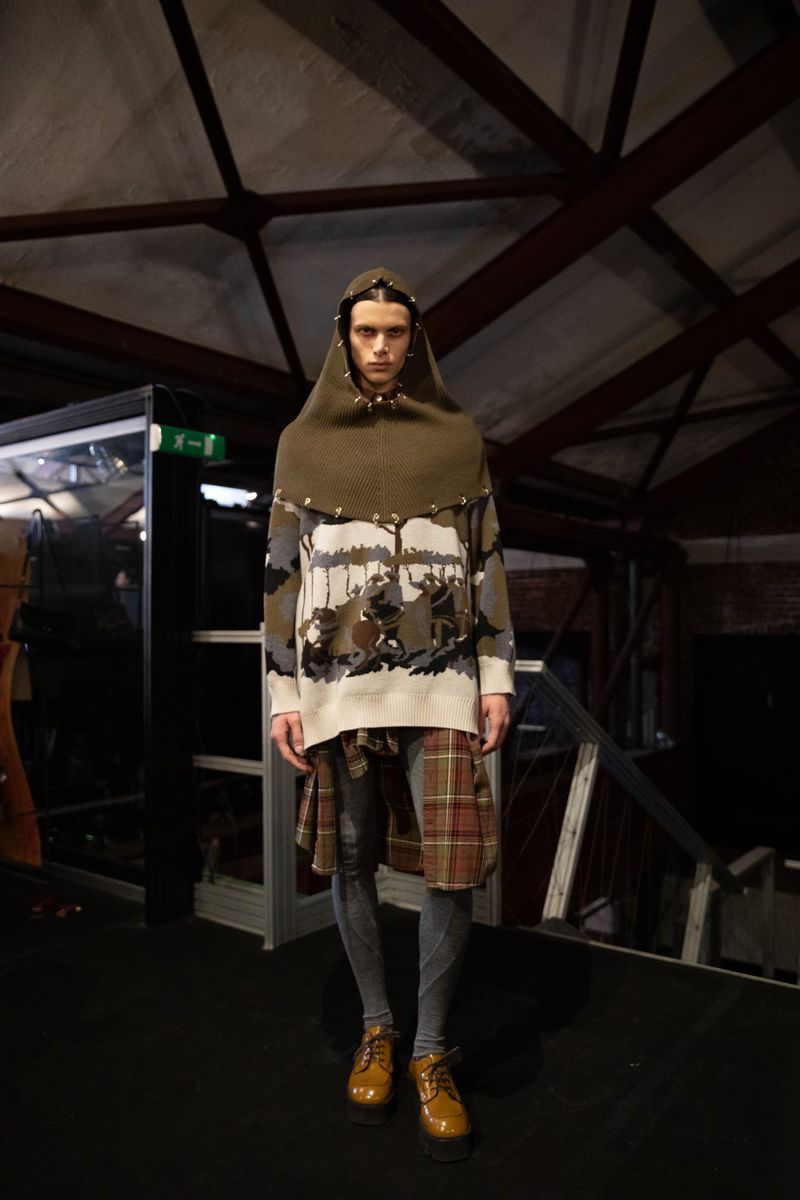 Male fashion model poses indoors wearing hooded sweater, plaid skirt over pants, and dress shoes in a dimly lit setting.