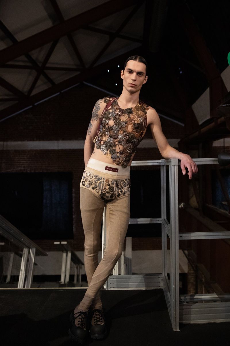 Androgynous fashion model poses in a decorative top, patterned bottoms, and leggings in a dimly lit, indoor location with industrial elements.