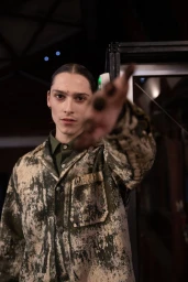 Model wearing a patterned jacket with an out-of-focus hand reaching towards the camera in a backstage setting.