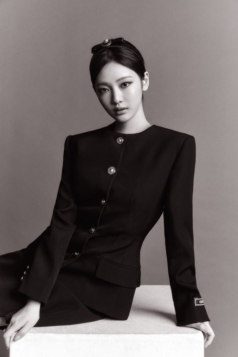 Monochrome portrait of a young Asian woman in a black dress, seated on a white block against a solid grey background, looking at camera.