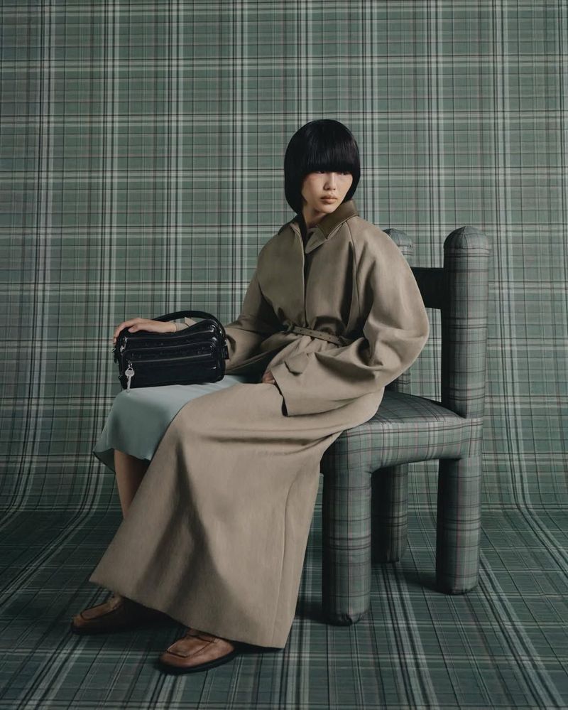 A woman with a blunt bob hairstyle sits on a plaid chair, wearing a trench coat, holding a handbag against a plaid background.