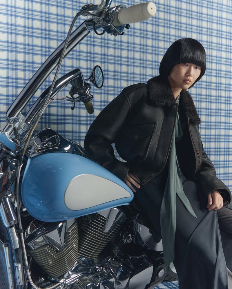 Stylish woman in a black aviator jacket sits on a blue and chrome motorcycle in front of a blue plaid background.