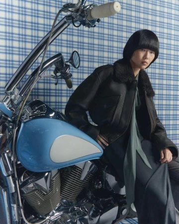 Stylish woman in a black aviator jacket sits on a blue and chrome motorcycle in front of a blue plaid background.