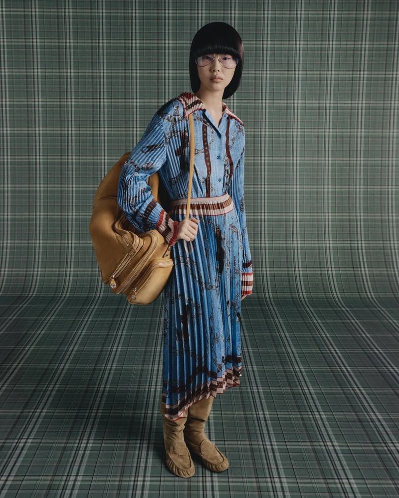 Fashion shot of an Asian woman in a patterned dress with a pleated skirt, wearing boots and glasses, and carrying a brown travel bag.