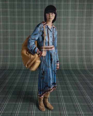 Fashion shot of an Asian woman in a patterned dress with a pleated skirt, wearing boots and glasses, and carrying a brown travel bag.