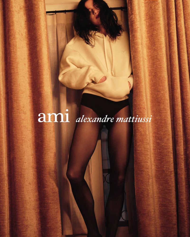 A fashion shot of a single model in a cream hoodie and black tights posing in front of gold curtains. Fashion photography, warm tones.