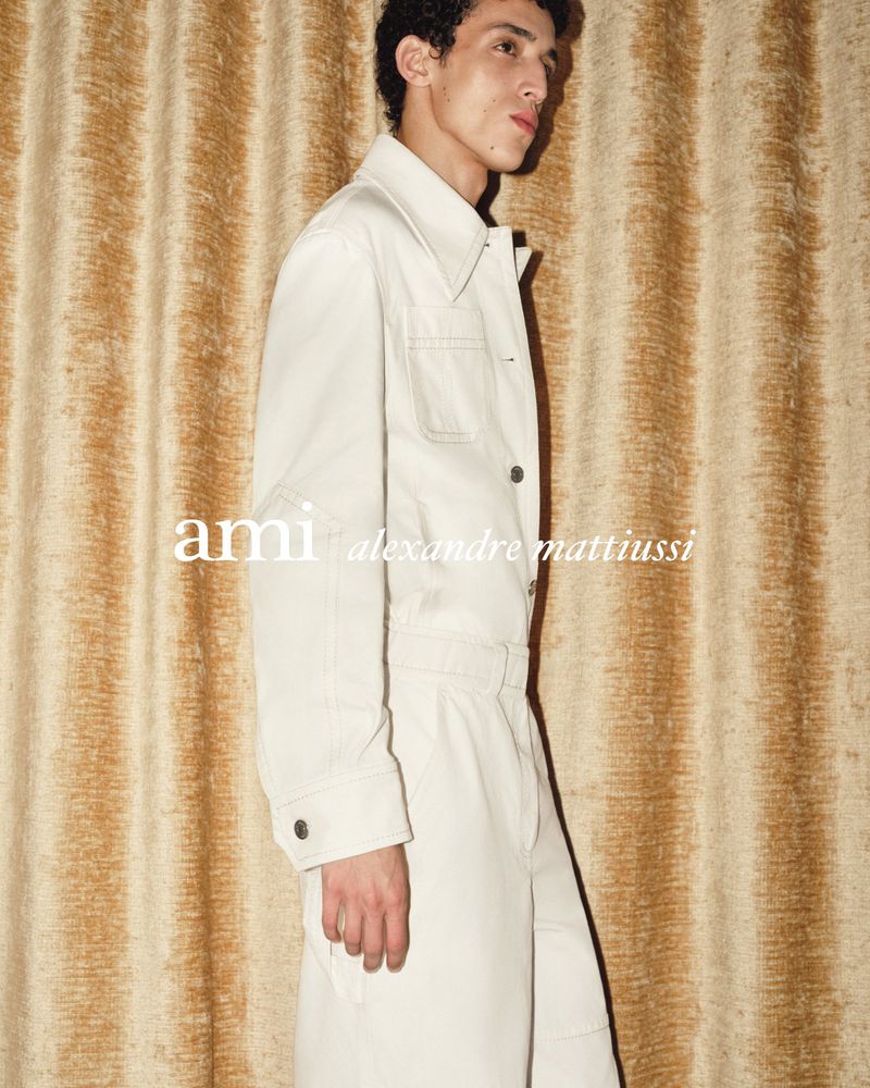 A young adult male model poses in a white collared jacket and pants against a textured gold backdrop in a fashion editorial shot.