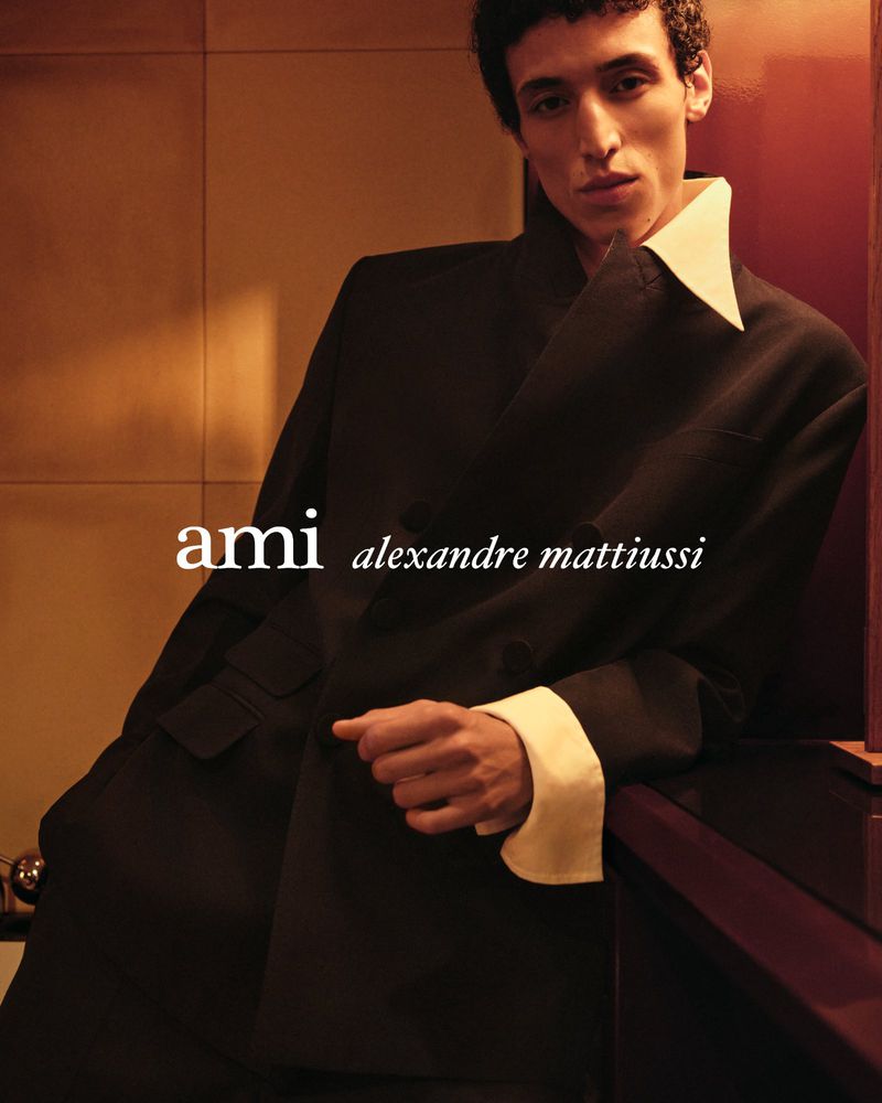 Fashion shot of a young adult male model in a black blazer and white shirt, with the brand name 'Ami' visible in the frame.