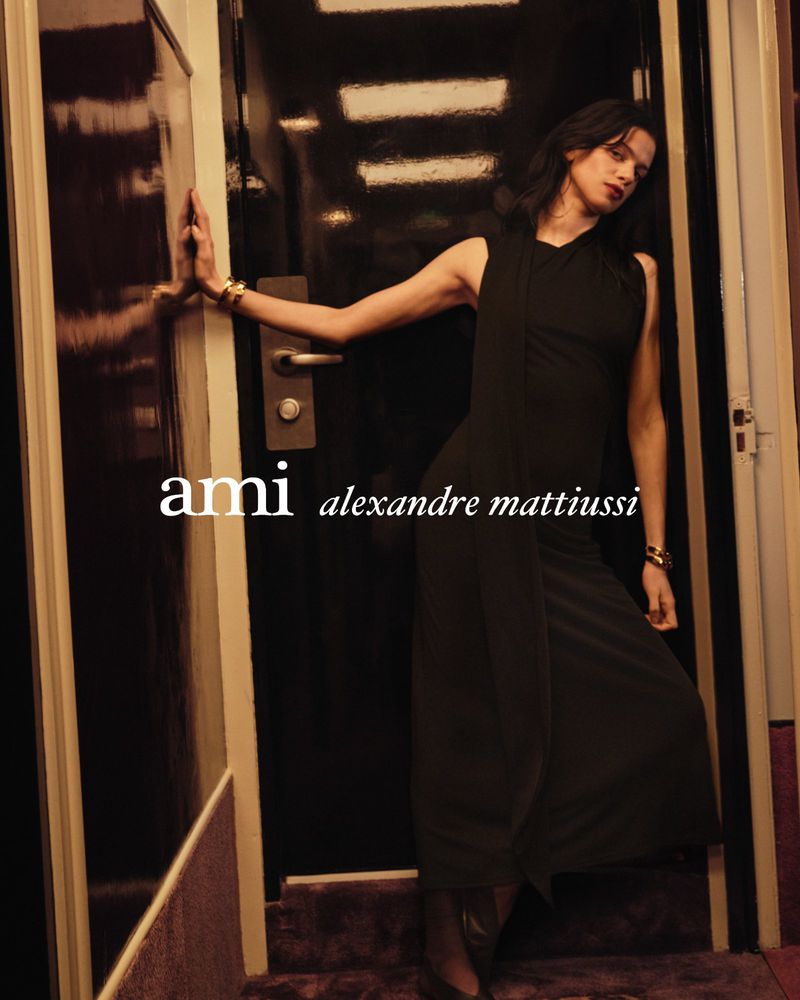 A woman in a black dress poses in a dimly lit corridor for an Ami Alexandre Mattiussi fashion campaign, wearing gold bracelets.