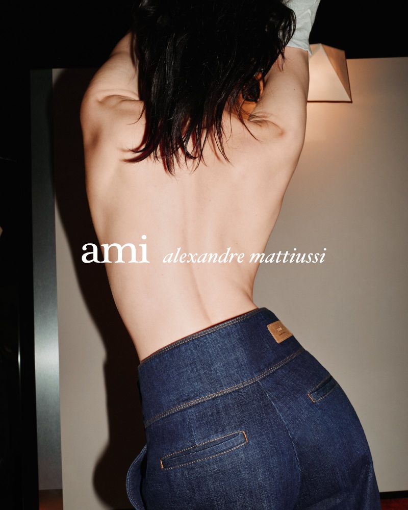 Woman with bare back posing in dark wash, high-waisted denim pants. Text: ami alexandre mattiussi. Interior fashion shot.