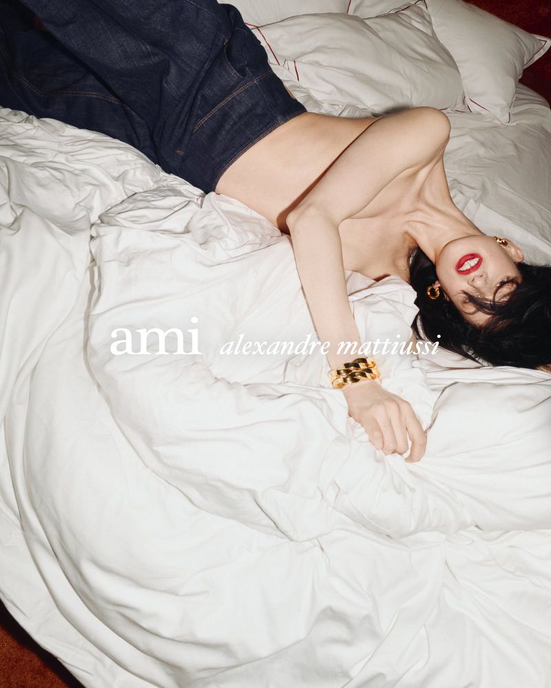 Woman lies on a white bed wearing dark jeans, a gold bracelet and earrings, and red lipstick with the brand name ami visible.