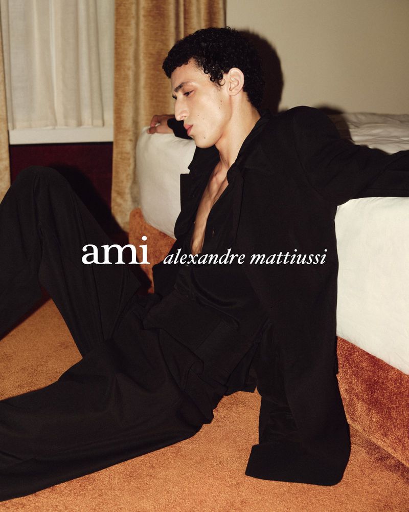 Fashionable young man posing on the floor wearing a black shirt and pants indoors near a white bed with beige walls, indoor lighting.