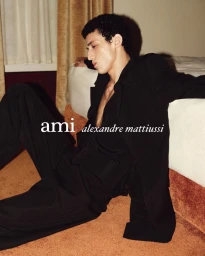 Male model wearing black attire leaning against a bed in an interior hotel room setting for Ami d hiver 26 campaign