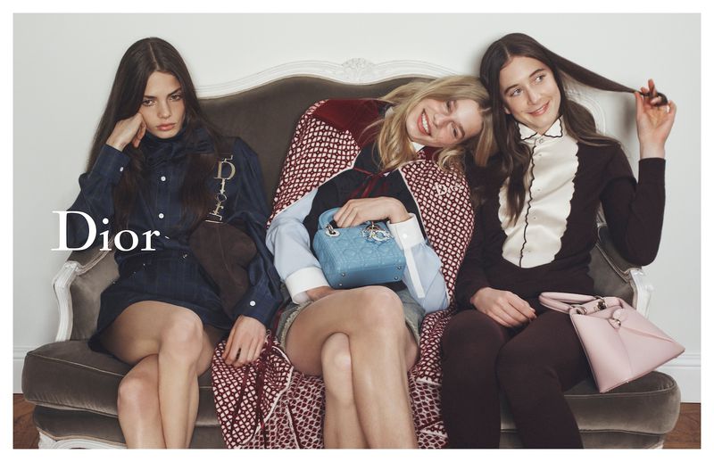 Three women wearing designer dresses and holding handbags pose on an ornate grey couch against a plain white wall in a fashion advertisement.