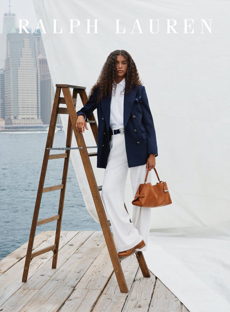 Fashion shot of a woman with curly hair wearing a blazer and standing on a ladder, holding a brown handbag, and buildings in the background.