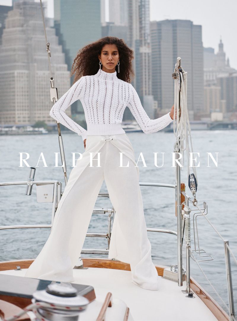 Elegant woman in white knit sweater and wide-leg pants poses on a yacht with an urban skyline backdrop in a fashion advertisement.