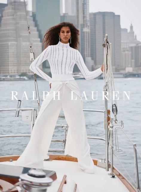 Elegant woman in white knit sweater and wide-leg pants poses on a yacht with an urban skyline backdrop in a fashion advertisement.