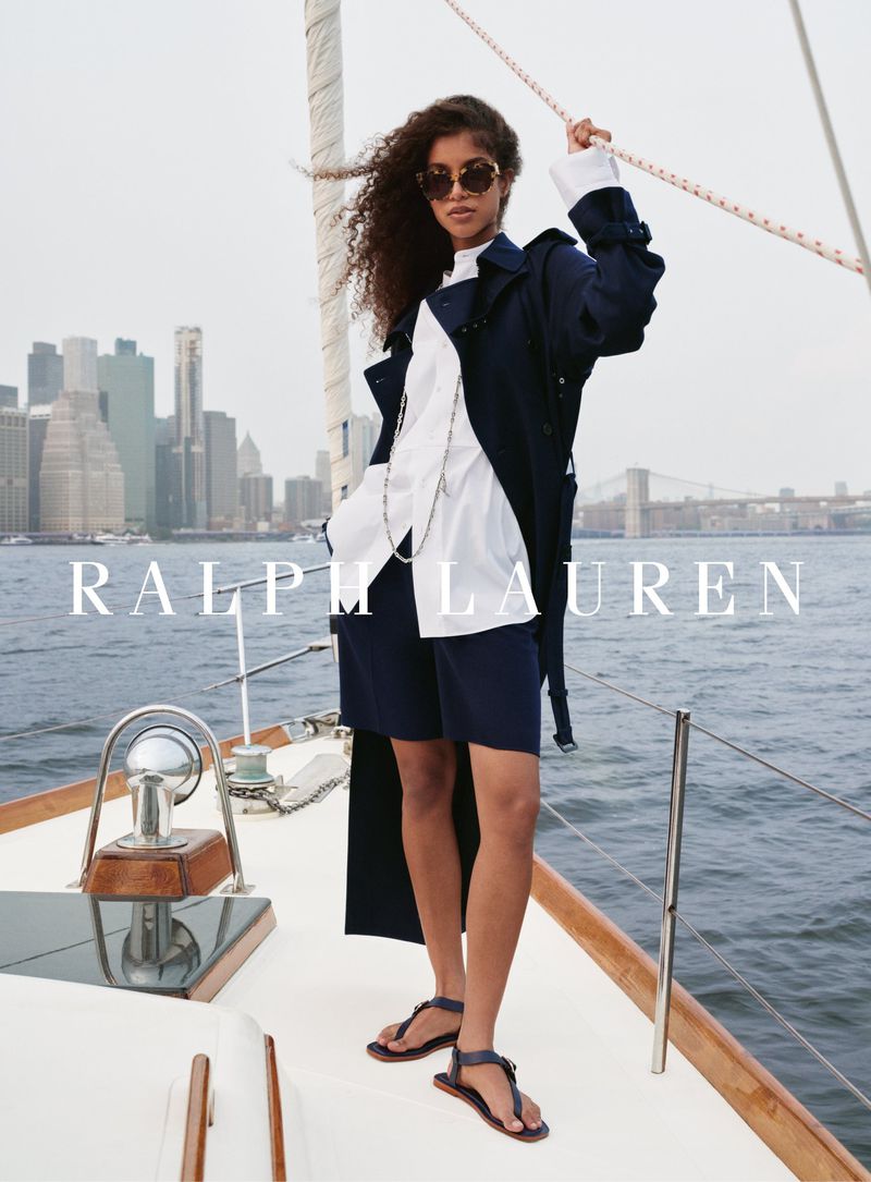 Brunette woman poses in a trench coat, button-down, and shorts on a yacht with a cityscape backdrop, creating a stylish nautical fashion image.