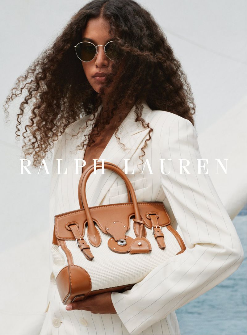 Fashion shot: Brunette model in striped jacket holding a tan leather handbag in front of a bright white and blue background.