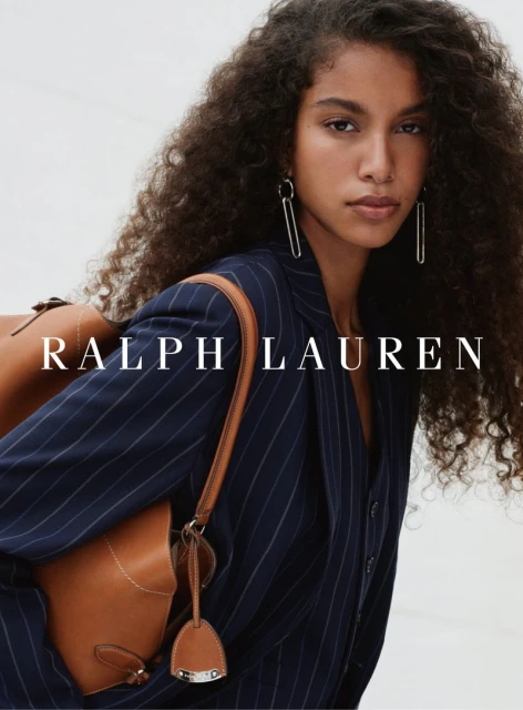Woman in pinstripe blazer carrying a brown leather satchel, posing in front of a neutral background with Ralph Lauren logo.
