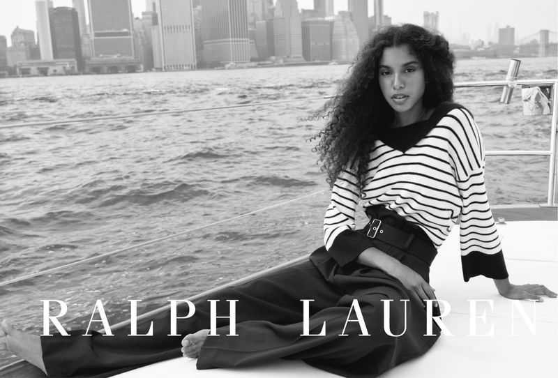 Black-and-white fashion shot of an adult female model wearing a striped long-sleeve shirt and wide-leg pants on a boat with a city skyline.