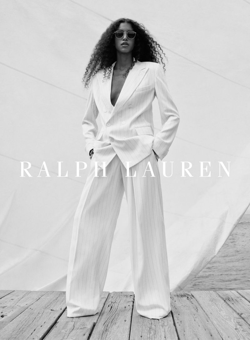 Fashion editorial shot of a Black female model wearing a white suit with sunglasses, posing against a sail on a wooden deck.