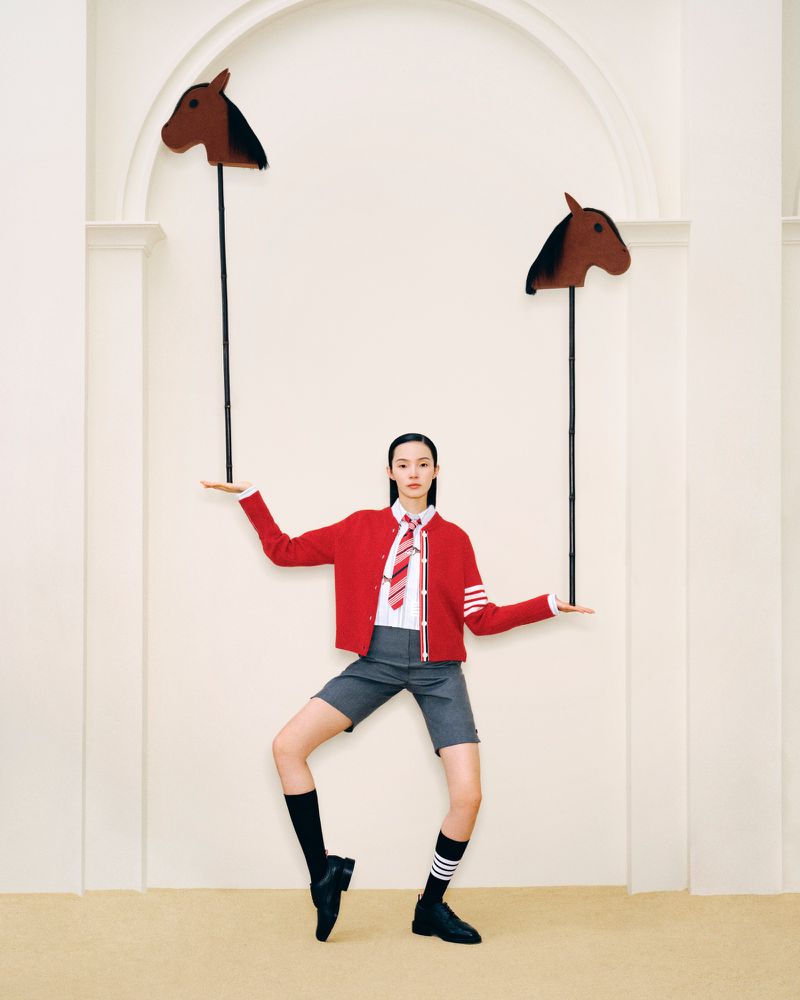 Stylish Asian woman in a red cardigan and grey shorts posing with whimsical horse head props in a minimalist photo studio setting.