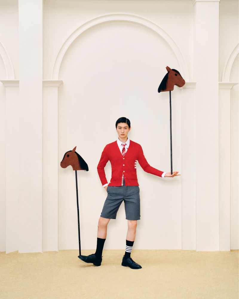 Stylish young man in red cardigan and gray shorts poses with toy horse sticks in an off-white studio setting with archways.
