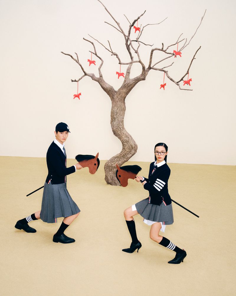 Two people face each other with toy horses in front of a tree with hanging horse decorations, set against a neutral-toned backdrop.