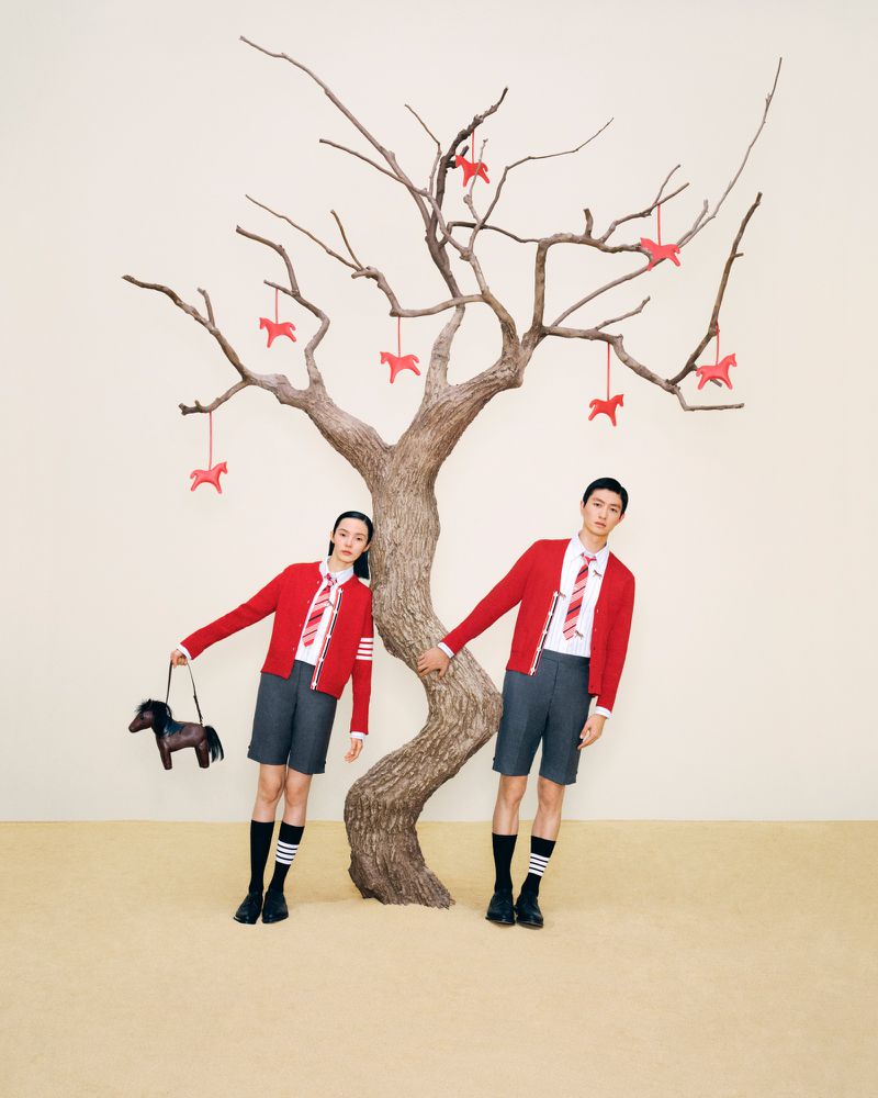 A fashionable couple poses in front of a barren tree adorned with horse ornaments. The pair wear matching school uniform-inspired outfits.