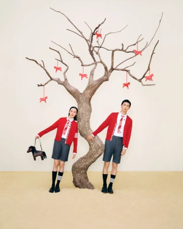 A fashionable couple poses in front of a barren tree adorned with horse ornaments. The pair wear matching school uniform-inspired outfits.