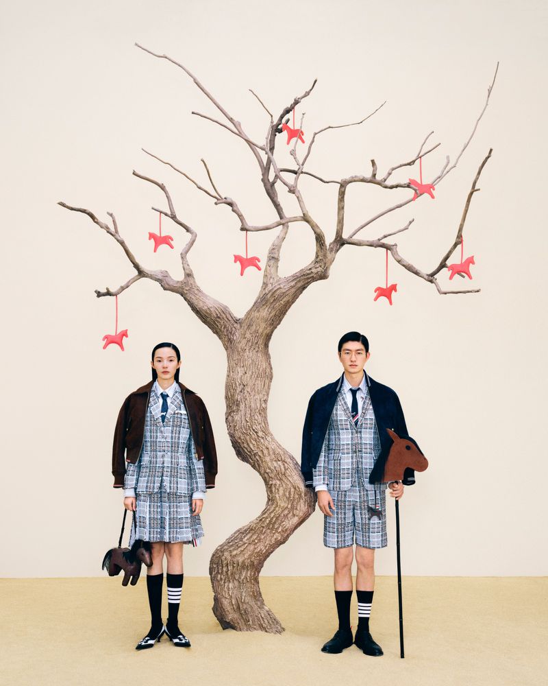 Two people in plaid suits standing near a bare tree adorned with horse figures, creating an editorial or fashion-focused composition.