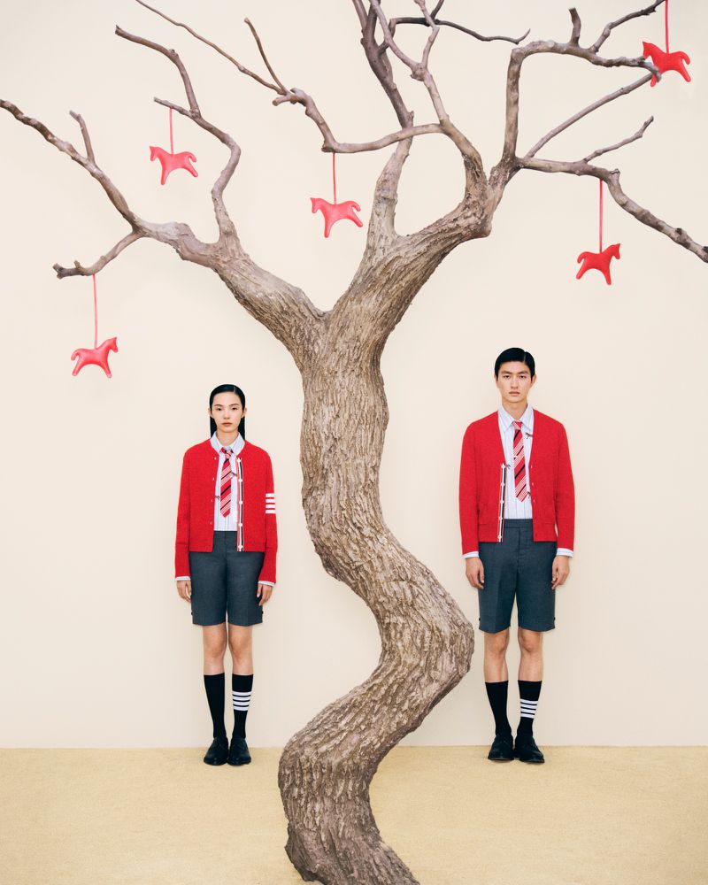 Two people in red cardigans and grey shorts pose in front of a bare tree with red horse ornaments in a minimal indoor setting.