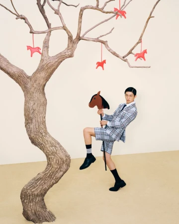 Jin Dachuan posing with a hobby horse in a Thom Browne plaid short suit next to a bare tree hung with red ornaments.