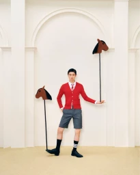 Model Jin Dachuan in a red Thom Browne cardigan and grey shorts posing with toy stick horses in an architectural white studio.