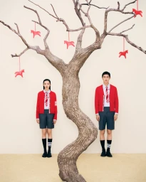 Models Xiao Wen Ju and Jin Dachuan in red cardigans and grey shorts standing beside a bare tree hung with red horse ornaments.