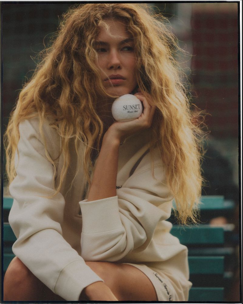A relaxed portrait of a young adult female with long wavy hair in casual attire, holding a white ball with text, outdoors on a bench.