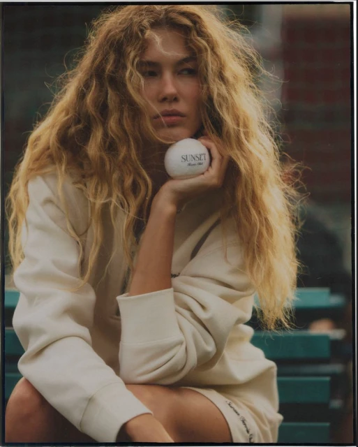 A relaxed portrait of a young adult female with long wavy hair in casual attire, holding a white ball with text, outdoors on a bench.