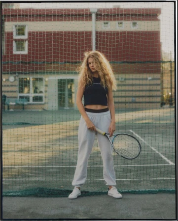 Female tennis player stands on a green court with a racket, wearing a black tank top, light sweatpants, and white sneakers in daylight.