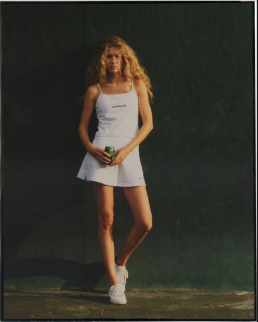 Woman in white tennis outfit poses against a green wall, holding a can. Blonde hair and white sneakers complete the look.