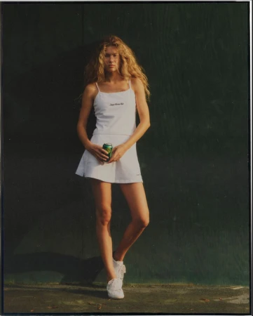 Woman in white tennis outfit poses against a green wall, holding a can. Blonde hair and white sneakers complete the look.