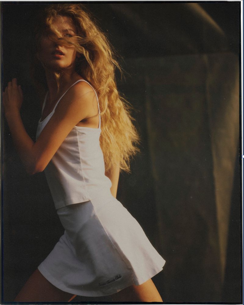 Woman with flowing blond hair posing in sunlight wearing a white tennis outfit against a dark backdrop in a studio environment.
