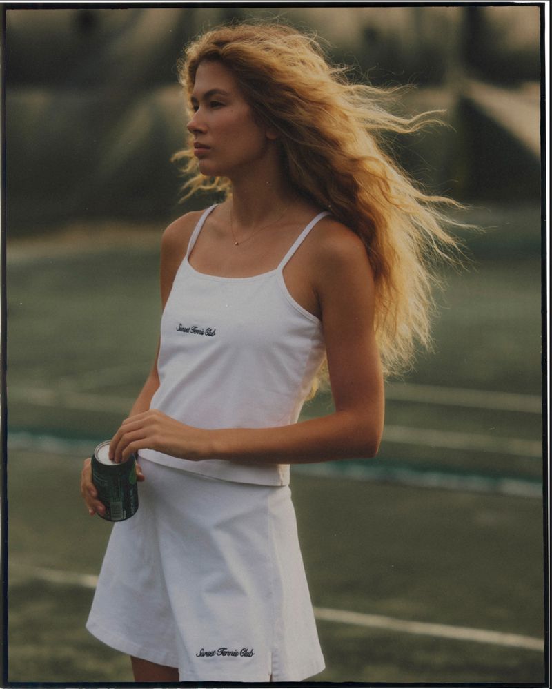 Young blonde woman on tennis court wearing white tennis dress holding a beverage can, sunny day, athletic style, outdoors