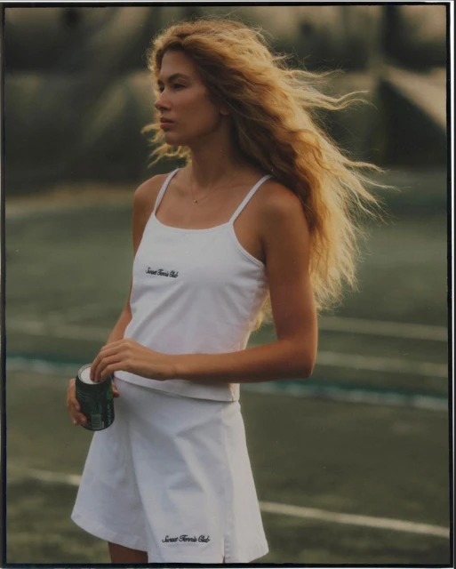 Young blonde woman on tennis court wearing white tennis dress holding a beverage can, sunny day, athletic style, outdoors